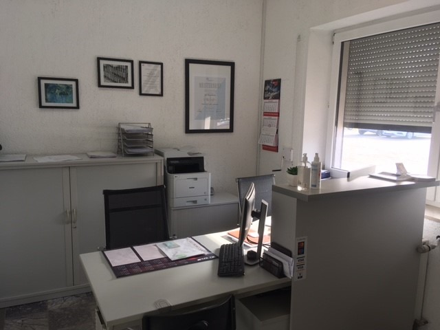 Wnętrze biura warsztatu samochodowego z biurkiem, komputerem, szafą na dokumenty i kalendarzem na ścianie. Widoczne certyfikaty w ramkach.