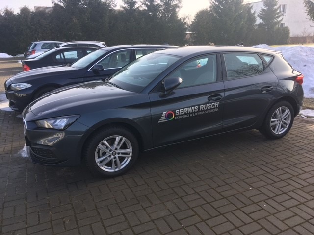 Szary samochód marki Seat z naklejonym logo serwisu 'Serwis Rusch. Eksperci w likwidacji szkód' na boku, zaparkowany na brukowanym parkingu w słoneczny dzień.