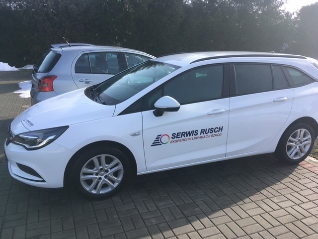 Biały samochód kombi z logo firmy 'Serwis Rusch - Eksperci w likwidacji szkód' zaparkowany na brukowanej powierzchni, z innym samochodem w tle.