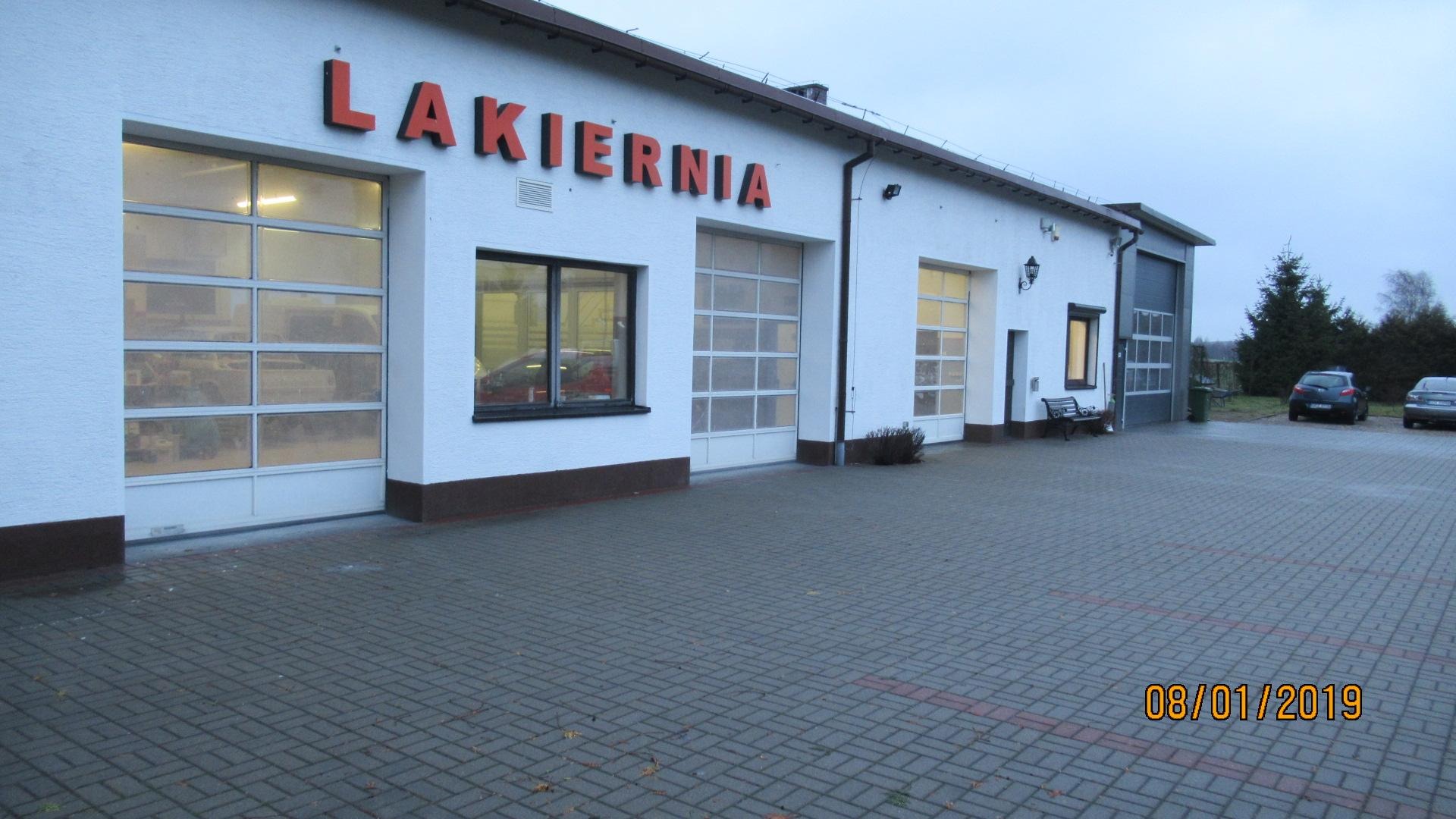 Budynek warsztatu z napisem 'LAKIERNIA' nad przeszklonymi bramami wjazdowymi, widoczny z zewnątrz na tle szarego nieba i kostki brukowej.