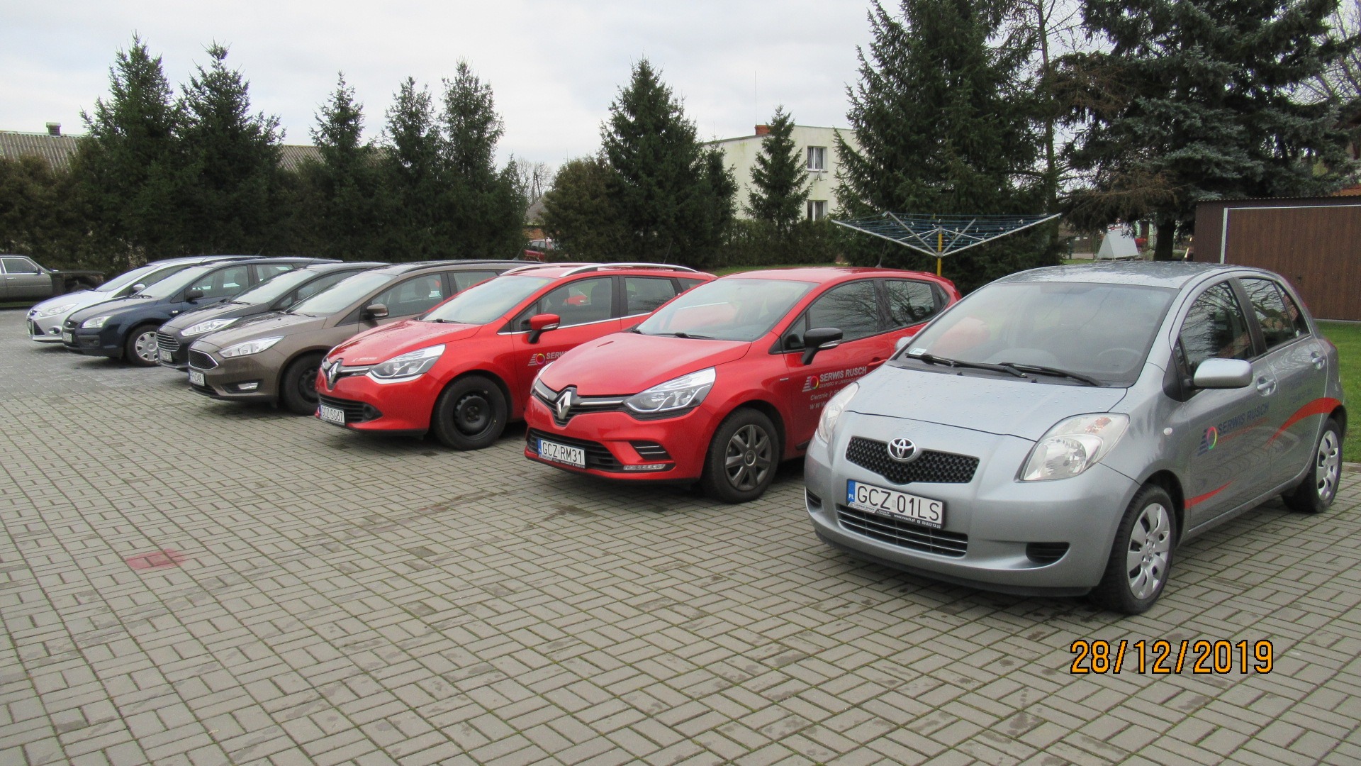 Rząd zaparkowanych samochodów różnych marek i kolorów na brukowanym parkingu przed budynkiem, z widocznym logo 'Serwis Rusch' na czerwonym Renault Clio i szarym Toyota Yaris.