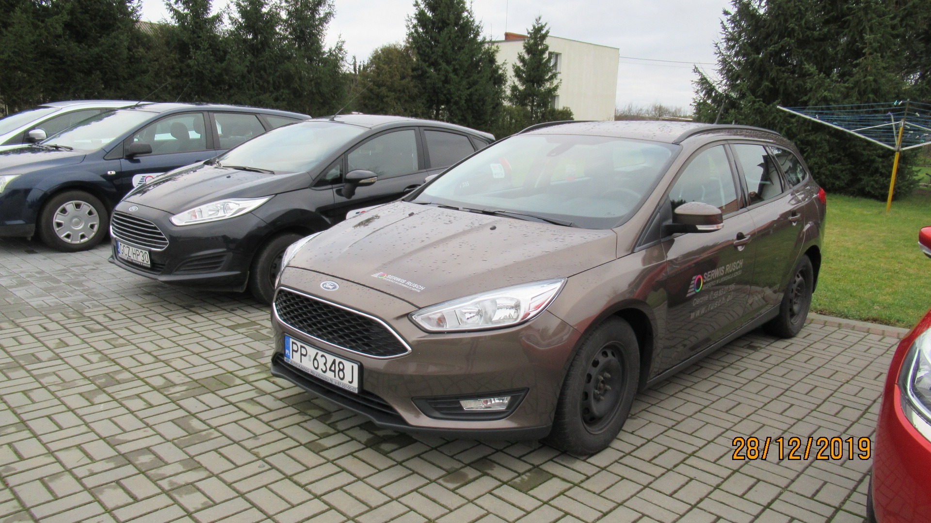 Brązowy Ford Focus kombi z naklejonym logo 'Serwis Rusch' na karoserii, zaparkowany na brukowanym placu obok innych samochodów Forda, w tle drzewa iglaste i budynek.