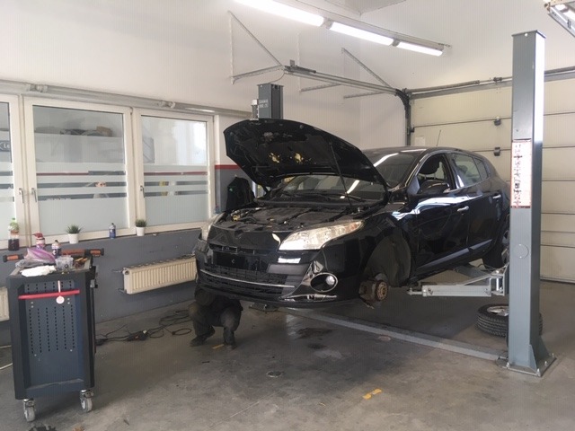 Czarny samochód na podnośniku w warsztacie, z otwartą maską i zdjętym kołem, widoczny mechanik pracujący pod autem.