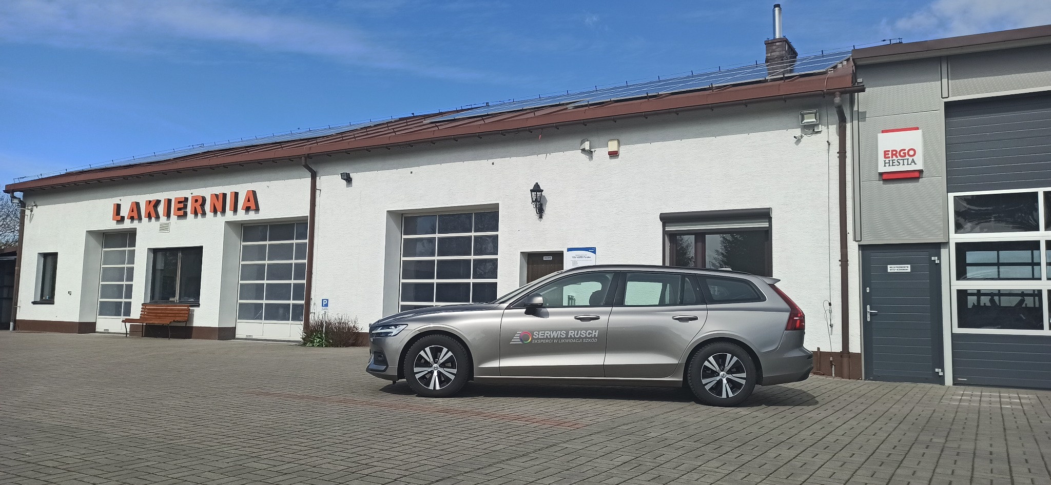 Szaro-srebrne kombi Volvo zaparkowane przed budynkiem warsztatu z napisem 'Lakiernia' i logo ERGO HESTIA, z widocznym oznaczeniem 'Serwis Rusek - Eksperci w likwidacji szkód' na boku auta.