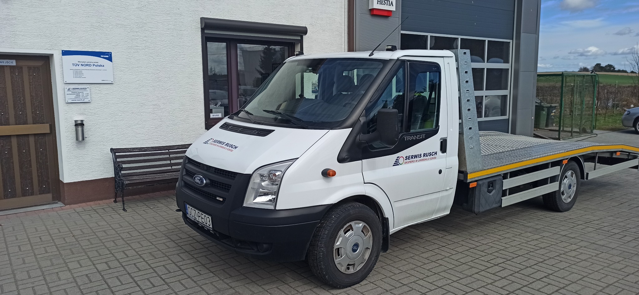 Biały Ford Transit z platformą lawetową zaparkowany przed budynkiem z szyldem TÜV NORD Polska i logo serwisu samochodowego Serwis Rusch