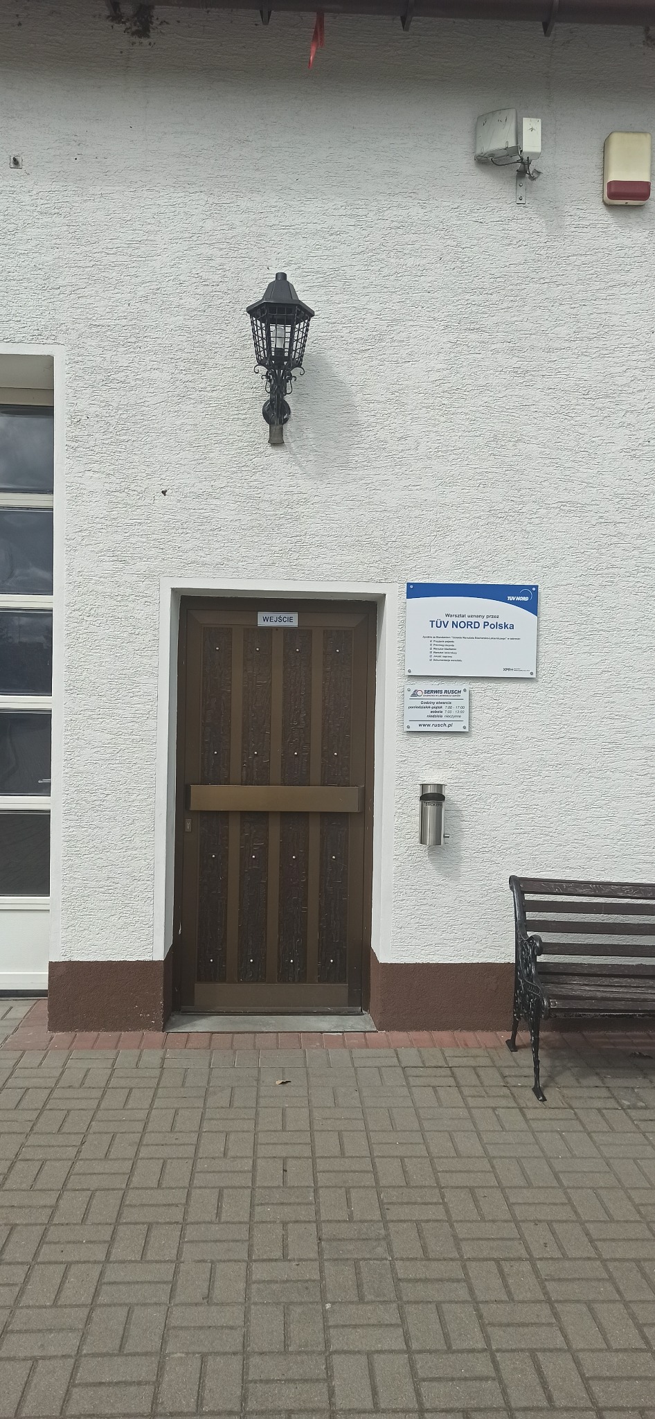 Wejście do warsztatu TUV Nord Polska, widoczna tablica informacyjna, lampa zewnętrzna i ławka.