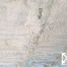 Świeżo nałożona, gruba warstwa białej pianki izolacyjnej PUR na suficie, z widocznymi metalowymi elementami mocującymi i logo firmy w prawym dolnym rogu.