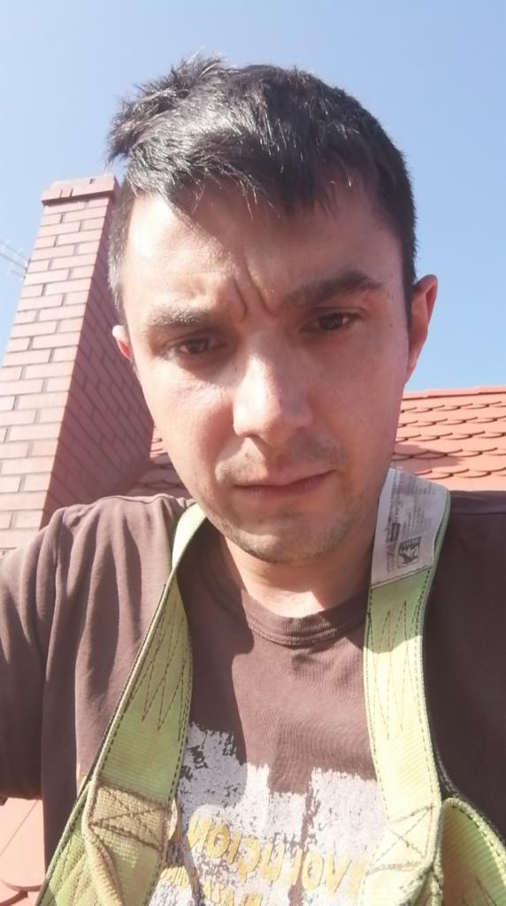 Mężczyzna w uprzęży na dachu z ceglaną konstrukcją w tle. Ujęcie z dołu, widok na fragment dachu z dachówką i komin. Jasne niebo w tle.