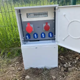 Otwarta skrzynka elektryczna z bezpiecznikami i gniazdami siłowymi (czerwone i niebieskie) na zewnątrz, obok metalowego ogrodzenia i trawy.