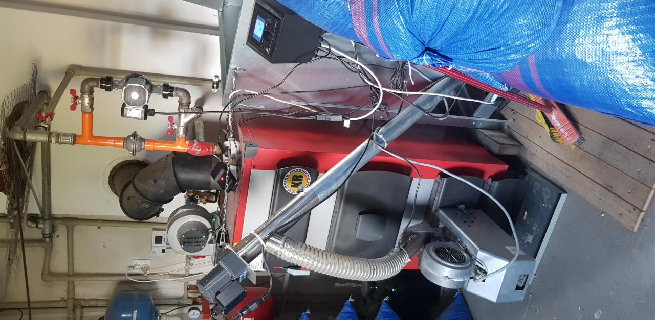 Czerwono-szary piec z podajnikiem, widoczne rury, zawory i elementy sterujące, niebieski worek w tle, widoczne okablowanie.