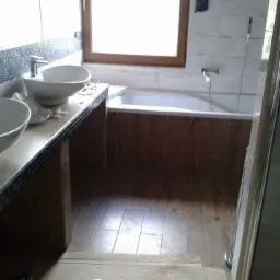 Wykończona łazienka z drewnopodobnymi płytkami na podłodze i obudowie wanny, dwie białe umywalki nablatowe z bateriami, okno z drewnianą ramą, białe płytki na ścianach.