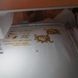 Proces wykonywania złotego haftu na białym materiale w ramce maszyny hafciarskiej; widoczne ozdobne motywy i inskrypcje.