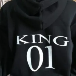 Czarna bluza z kapturem, widok z tyłu, z białym nadrukiem 'KING 01', w tle wieszaki z kolorowymi koszulkami.