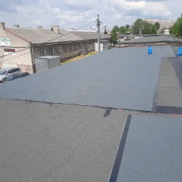 Fragment płaskiego dachu pokrytego nową papą termozgrzewalną, widoczne butle gazowe i ślady po zgrzewaniu, w tle budynki przemysłowe i drzewa.