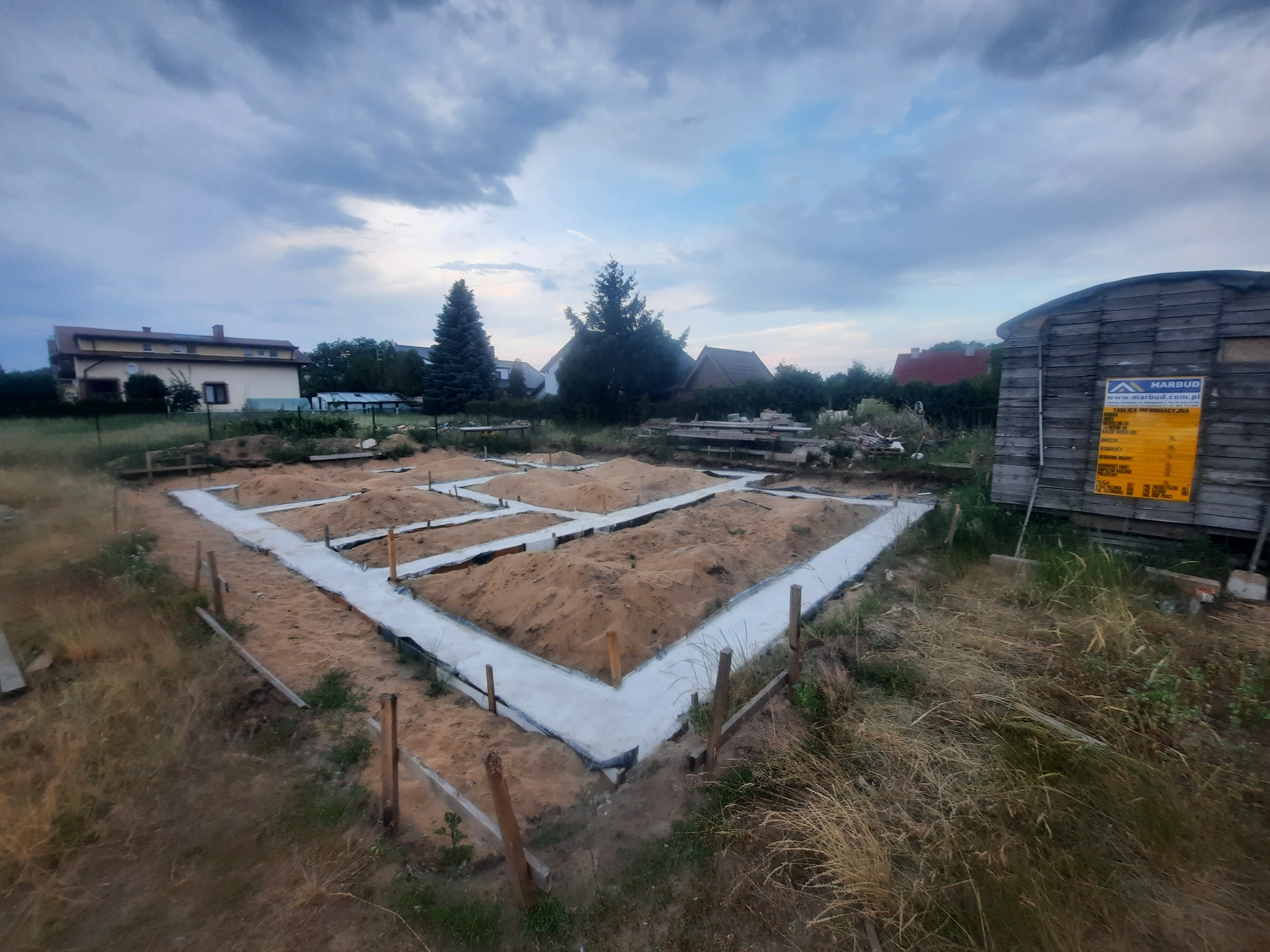Wylane fundamenty pod dom jednorodzinny z widoczną izolacją i szalunkiem, na tle zabudowań mieszkalnych i zachmurzonego nieba.