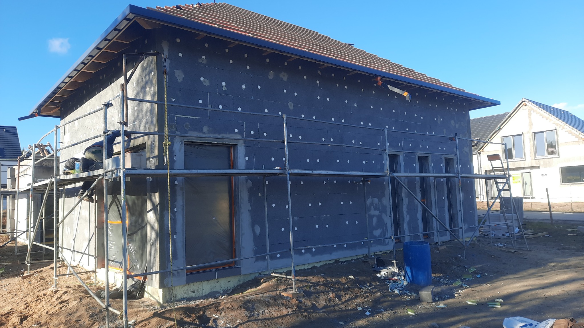 Budynek w trakcie ocieplania, widoczne rusztowania, pracownik na rusztowaniu, ściany pokryte szarym materiałem izolacyjnym z białymi kołkami, fragment dachu z dachówką, błękitne niebo w tle.