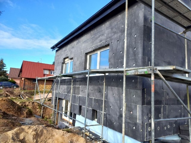 Dom w trakcie ocieplania czarnym styropianem, widoczne rusztowania i wykop pod fundamenty, sąsiednie domy w tle.