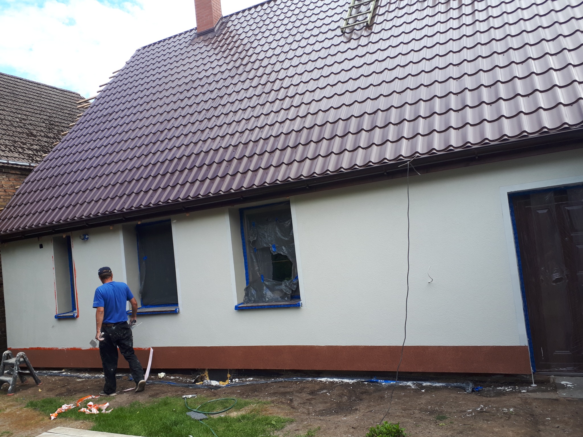 Pracownik w niebieskiej koszulce przygotowuje elewację domu z nowym, brązowym dachem z blachodachówki, okna zabezpieczone folią, widoczny fragment drabiny na dachu.