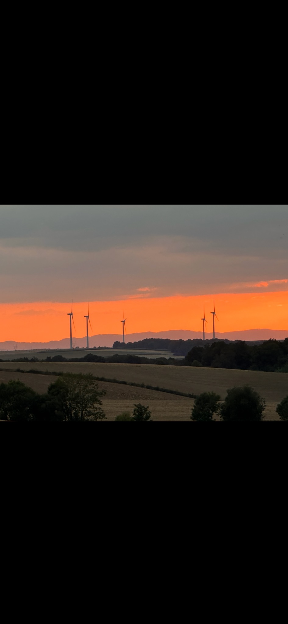 Krajobraz z turbinami wiatrowymi na wzgórzach o zmierzchu, pomarańczowe niebo, pola i lasy w oddali. Widok z perspektywy.