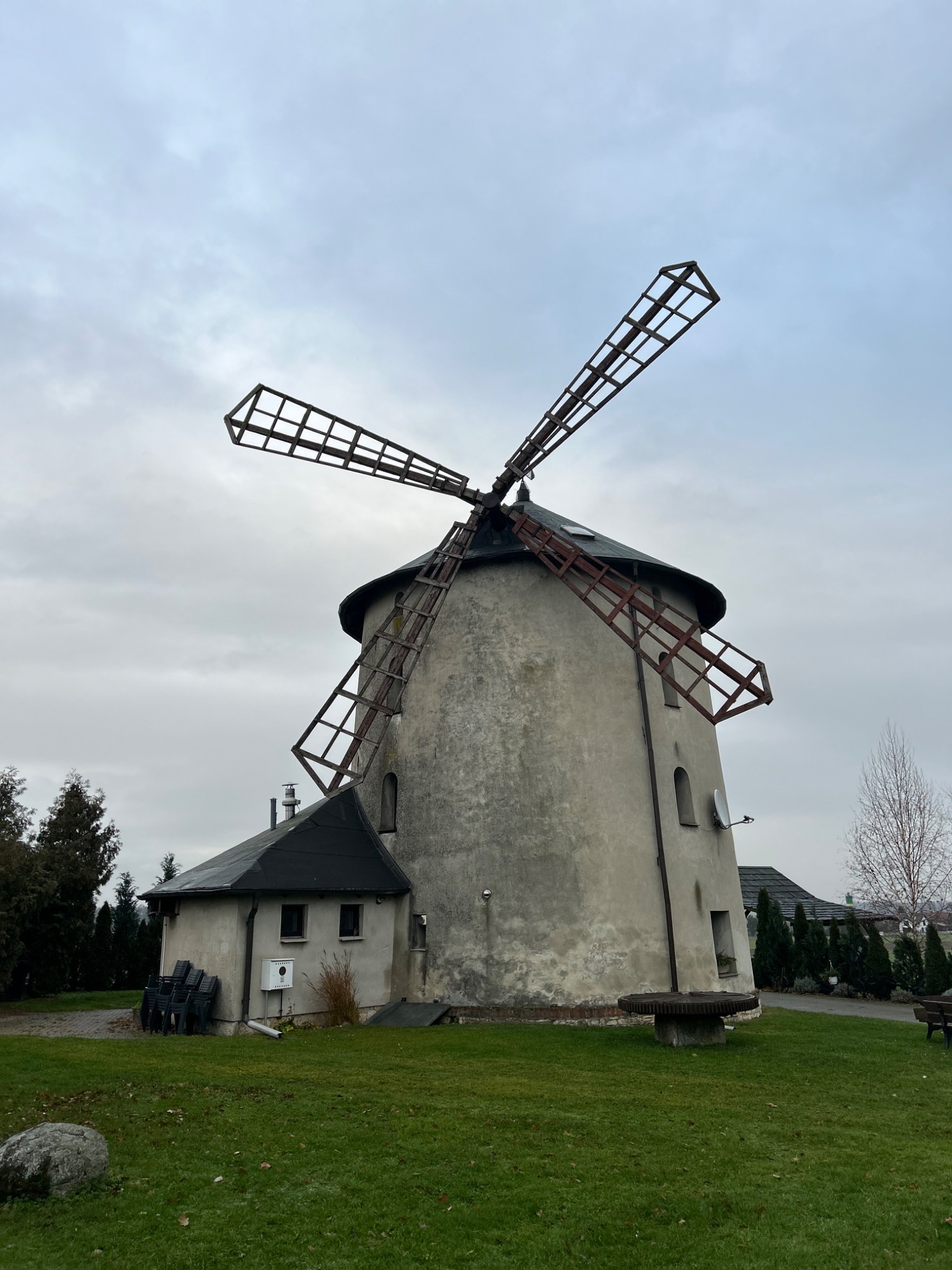 Stary, kamienny wiatrak z drewnianymi skrzydłami na tle pochmurnego nieba, otoczony zielonym trawnikiem i niskimi drzewami. Widoczny budynek przylegający do wiatraka.