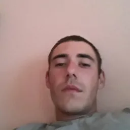 Portret mężczyzny z krótkimi, ciemnymi włosami na tle jednolitej, jasnoróżowej ściany. Ujęcie z dołu, widoczna część ramion w szarej koszulce.