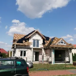 Dom w stanie surowym otwartym z częściowo położoną dachówką bitumiczną na drewnianej konstrukcji dachu, widoczne rusztowania i samochody w tle.