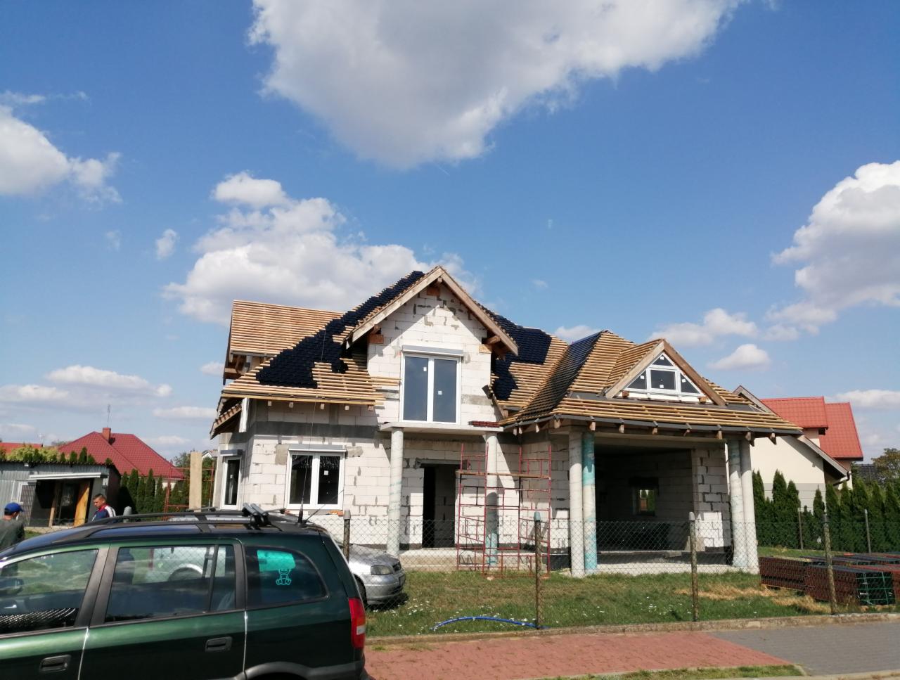 Dom w stanie surowym otwartym z częściowo położoną dachówką bitumiczną na drewnianej konstrukcji dachu, widoczne rusztowania i samochody w tle.