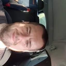 Portret uśmiechniętego mężczyzny z brodą w samochodzie, siedzącego na fotelu pasażera, z widocznym fotelikiem dziecięcym z tyłu. Zdjęcie obrócone.