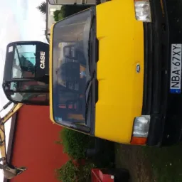 Żółty samochód Ford Transit z widoczną kabiną koparki CASE na przyczepie, zaparkowany na trawniku przed czerwoną ścianą budynku.