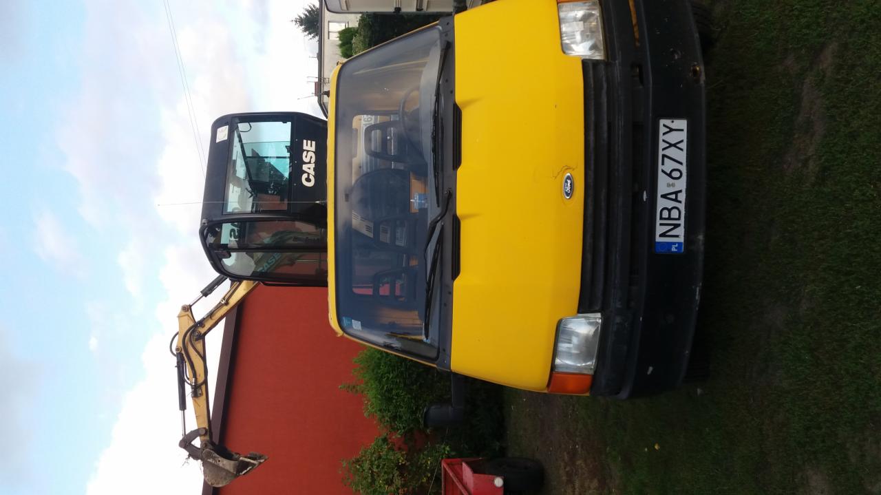 Żółty samochód Ford Transit z widoczną kabiną koparki CASE na przyczepie, zaparkowany na trawniku przed czerwoną ścianą budynku.