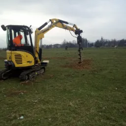 Żółta minikoparka Yanmar VIO27 z wiertnicą hydrauliczną wiercąca dziurę w zielonym trawiastym polu z pasącymi się końmi w tle, pochmurny dzień.