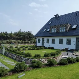 Biały dom z ciemnym dachem i zielonymi okiennicami, otoczony zadbanym trawnikiem z ozdobnymi krzewami i kamiennym murkiem oporowym, widok z poziomu trawnika.