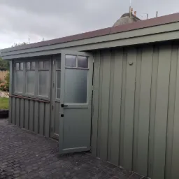 Szara, drewniana dobudówka z oknami i otwartymi drzwiami, na tle brukowanej nawierzchni i ogrodu z kamiennym murkiem.