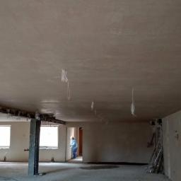 Artur Głowacz - Świeżo położone tynki maszynowe na suficie w przestronnym, jasnym pomieszczeniu w trakcie remontu, z widocznymi przewodami i elementami konstrukcyjnymi.