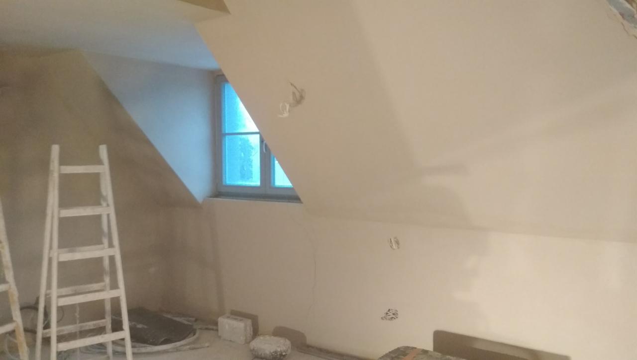 Poddasze ze świeżo nałożonymi gładziami, widoczne ślady po przewodach elektrycznych, drabina malarska i okno dachowe w tle.