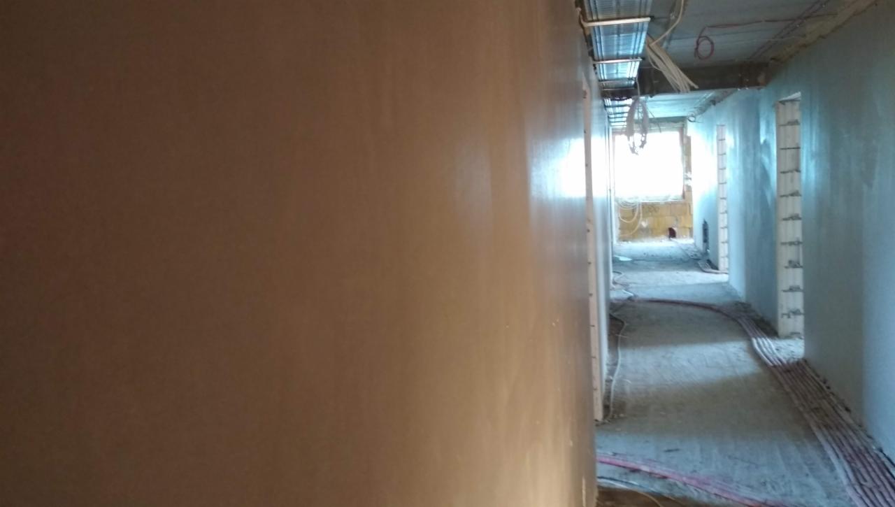 Świeżo położony, gładki tynk na ścianie w korytarzu podczas remontu, widoczne instalacje pod sufitem i przewody elektryczne na podłodze.