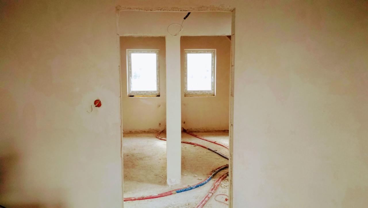 Świeżo otynkowane ściany w pomieszczeniu z dwoma oknami, widoczne kable instalacji podłogowej, surowy stan wykończenia.