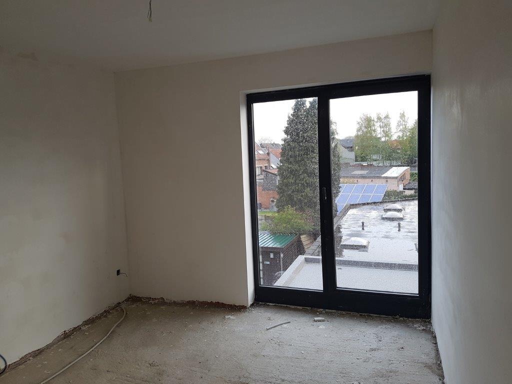 Wnętrze pokoju w stanie surowym po nałożeniu gładzi gipsowej na ścianach, widoczne okno balkonowe z czarną ramą i widok na zabudowania z panelami słonecznymi.