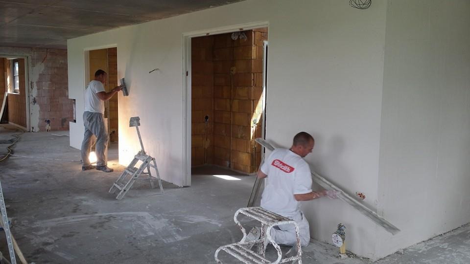 Dwóch mężczyzn w trakcie nakładania tynku na ściany w surowym wnętrzu, widoczne narzędzia i drabiny malarskie, w tle ceglany mur w otworze drzwiowym.