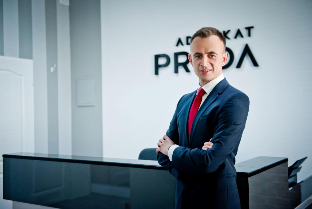 Portret uśmiechniętego mężczyzny w granatowym garniturze i czerwonym krawacie, stojącego na tle minimalistycznej recepcji z napisem 'ADWOKAT PRO...', skrzyżowane ręce, profesjonalny wygląd.
