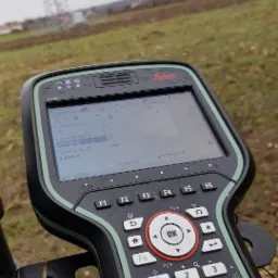 Geodezyjny pomiar wysokości anteny 2.0000 m za pomocą odbiornika GPS Leica na tle pola i wieży telekomunikacyjnej w pochmurny dzień.