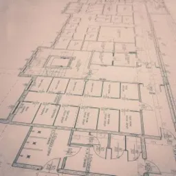 Szkic architektoniczny rzutu poziomego budynku z zaznaczonymi pomieszczeniami, wymiarami i elementami konstrukcyjnymi, wykonany w odcieniach sepii.