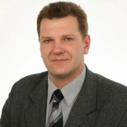 Portret mężczyzny w garniturze, Zawady. Uśmiechnięta twarz, jasne tło. Widoczna przypinka w klapie marynarki i wzorzysty krawat.