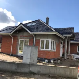 Dom w trakcie budowy: widoczna surowa elewacja z czerwonej cegły, zamontowane białe okna PCV, czarny dach z dachówki ceramicznej oraz białe rynny, na pierwszym planie betonowy fundament.