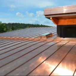 Miedziany dach z wyraźnie widocznymi panelami i świetlikiem, z drewnianą nadbudówką okienną na tle zieleni drzew i błękitnego nieba.