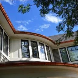 Elegancki dom z widocznymi miedzianymi obr&oacute;bkami blacharskimi dachu i rynnami, na tle błękitnego nieba z chmurami i zieleni drzew.