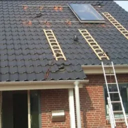 Dom z ciemną dachówką, na której widoczne są drewniane drabinki dachowe i drabiny przystawne, sugerujące prace naprawcze lub montaż instalacji solarnych.