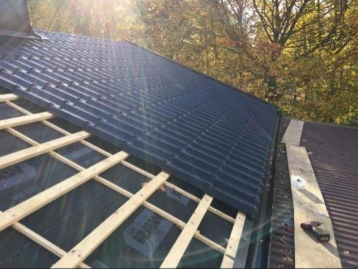 Częściowo pokryty, ciemny dach z blachodachówki, widoczne elementy więźby dachowej i narzędzie pracy, w tle drzewa.