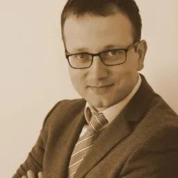 Portret mężczyzny w garniturze i okularach, z subtelnym uśmiechem, na jednolitym, kremowym tle, utrzymany w sepii.