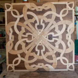 Panel dekoracyjny z drewna w dwóch odcieniach, ułożony w skomplikowany, symetryczny wzór przypominający przeplatające się wstęgi i serca, na tle warsztatu z narzędziami i półkami.